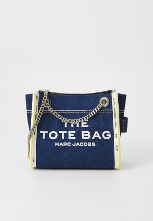 Denim tote bag i mørkeblå med hvid tekst, sølv kædetras, og gule accenter. Har en struktureret form og åben topdesign.