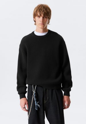 Young person with light brown hair wearing a black sweater over white shirt, black pants, and chain key accessory hanging from waist.