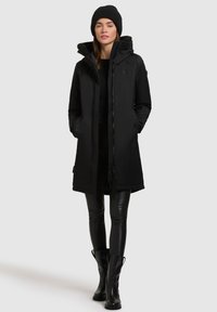 Parka neagră impermeabilă cu glugă, dotată cu fermoar, două buzunare frontale și interior căptușit cu fleece, purtată cu colanți negri și bocanci.