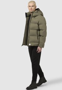 Chaqueta acolchada verde oliva con capucha, presenta detalles en negro, bolsillos con cremallera y un parche con el logo, combinada con pantalones y botas negras.