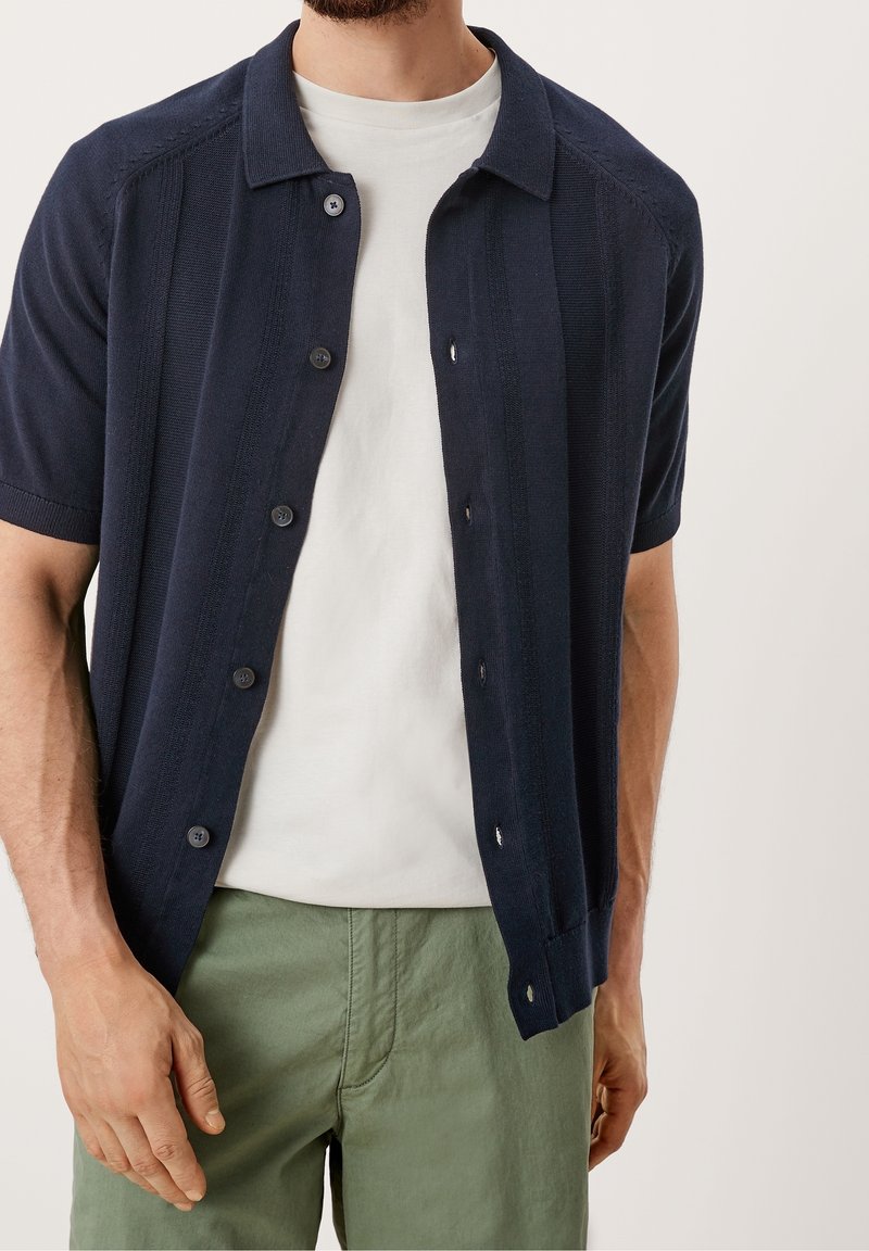 Marineblauw kortoe shirt met knopen, gemaakt van gestructureerde stof, met een kraag, verticale strepen en donkere knopen, gecombineerd met lichtgroene broeken.