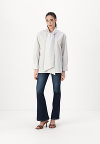 Blouse rayée gris clair avec un col en écharpe noué, associée à un jean flare noir et des sandales à talons ornées de clous.