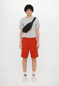 Černá crossbody taška s elegantním designem, šedé tričko, červené šortky a béžové tenisky; taška má nastavitelné rameno a detaily loga.