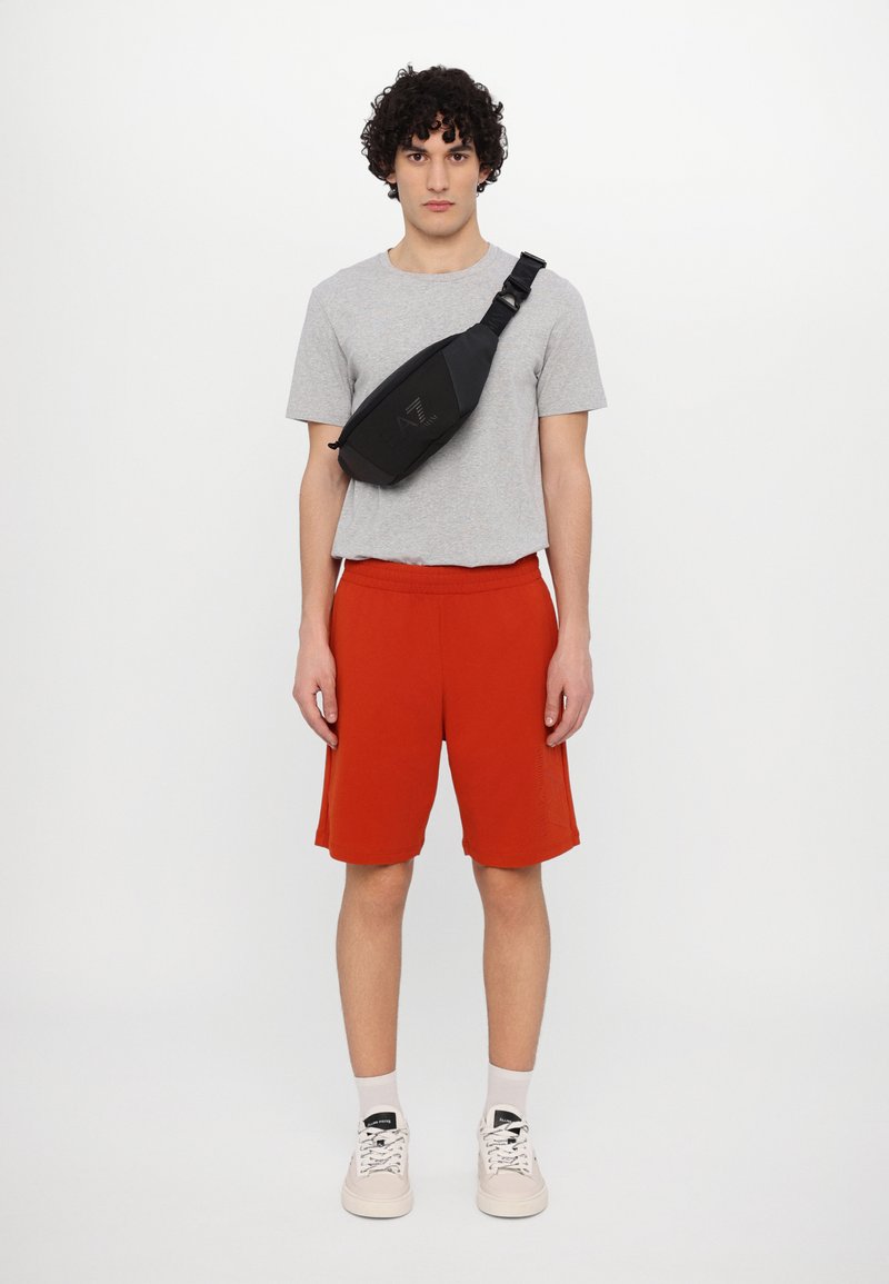 Černá crossbody taška s elegantním designem, šedé tričko, červené šortky a béžové tenisky; taška má nastavitelné rameno a detaily loga.
