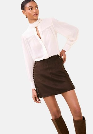 Weiße Bluse mit plisseeartiger Verzierung und Schlitz am Ausschnitt, kombiniert mit einem braunen Wildleder-Minirock mit Ösenakzenten an den Seiten.