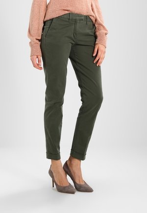 Vrouw draagt olijfgroene slim-fit broek, perzikkleurige gebreide trui en grijze suède hoge hakken, staand tegen een effen lichte achtergrond.