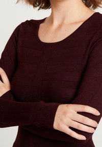 Haut bordeaux à manches longues avec un col rond, présentant des rayures horizontales texturées. Confectionné en maille douce avec des poignets côtelés.