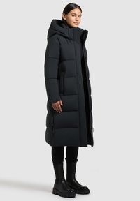 Lange zwarte pufferjas met capuchon, met een gestikte uitstraling, twee zijzakken en een slanke silhouet. Draagt over zwarte kleding en laarzen.