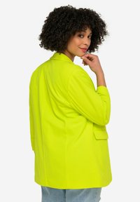 Blazer vert néon avec une coupe ajustée, fermeture à un bouton et revers crantés. Il présente une poche latérale et des épaulettes structurées.