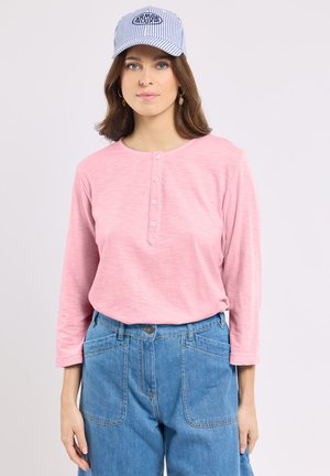 Jeune femme portant une casquette rayée bleu et blanc, une chemise rose à manches longues boutonnée, un jean bleu taille haute et des boucles d'oreilles dorées en forme d'ananas.