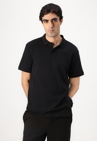 Zwarte poloshirt van textuurstof, met een kraag en een drievoudige knopenlijst. Draagt de handen in de zakken, in combinatie met zwarte broek.