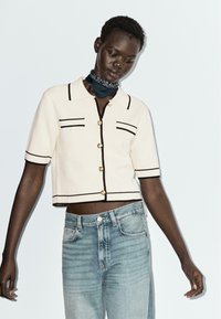 Polo corto a maniche corte color crema con rifiniture nere e dettagli con bottoni, abbinato a jeans a vita alta blu chiaro. Gli accessori includono una sciarpa scura.