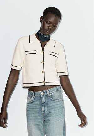 Polo cropped crème à manches courtes avec des détails noirs et de boutons, associé à un jean taille haute bleu clair. Les accessoires comprennent une écharpe sombre.