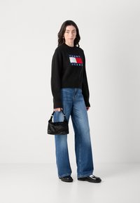 Svart strikket genser med "TOMMY JEANS"-logo, samparet med lyseblå flarejeans. Holder en liten svart håndveske med teksturert overflate.