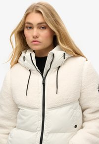 Doudoune blanche avec un haut en molleton doux et texturé, fermeture éclair, capuche avec cordon de serrage et accents noirs. Convient pour les temps froids.
