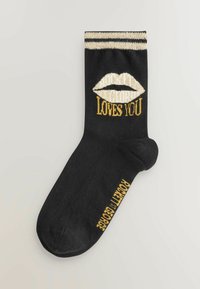 Schwarze Socke mit goldenen und weißen Texten, auf denen "Liebt Dich" steht, sowie einem Lippen-Graphic. Mit einem gestreiften goldenen Bund. Hergestellt aus einem glatten Stoff.