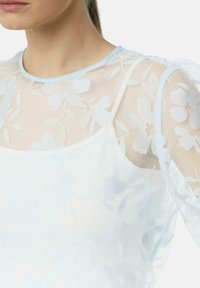 Helleblaue, blütenförmige, transparente Bluse mit Puffärmeln, die ein zartes Blumenmuster und einen runden Ausschnitt aufweist, über einer einfarbigen Basis geschichtet.