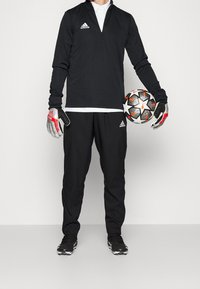 Schwarzes Sportoutfit mit einem langärmeligen Reißverschluss-Shirt und Hose, dazu schwarze Handschuhe und einen Fußbal. Die Handschuhe haben rote Akzente.