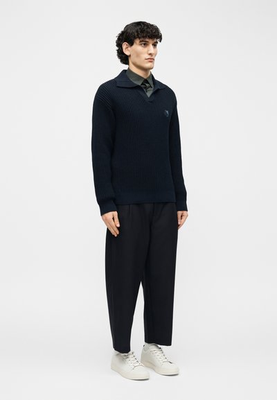 Jeune homme aux cheveux bouclés foncés portant un pull en laine foncé, un pantalon noir et des baskets blanches, debout devant un fond blanc uni.