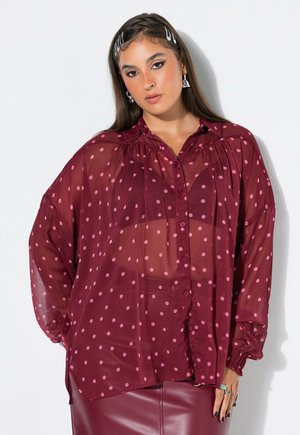 Donna che indossa una camicetta trasparente bordeaux a pois rosa, gonna in pelle bordeaux abbinata e orecchini geometrici argento su sfondo bianco.