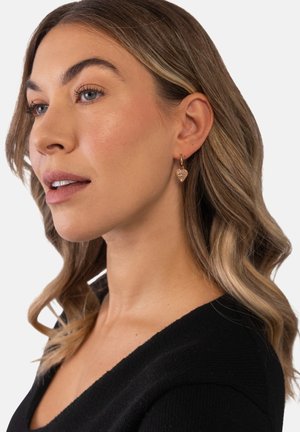 Frau mit welligem blondem Haar, die herzförmige, hängende Ohrringe trägt und ein schwarzes Oberteil anhat, schaut leicht zur Seite vor einem schlichten Hintergrund.