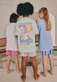 Tre barn står med ryggen mot kameran, iklädda färgglada, oversized T-shirts och mönstrade shorts. Utmärkande mönster inkluderar blommotiv.