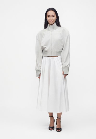 Femme aux longs cheveux foncés portant une veste zippée gris clair avec de larges manches et un col haut, une jupe midi blanche et des sandales noires à talons hauts.