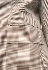 Poche à rabat et détail passepoilé sur un blazer à motif pied-de-poule marron et blanc en tissu texturé.