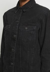 Acercamiento de una persona que lleva una chaqueta de mezclilla negra con bolsillos en el pecho de solapa abotonados y detalles de costura visibles.