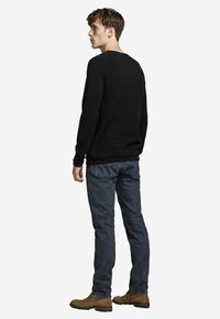 Črna dolga rokavica, slim fit, okrogla izrez. V kombinaciji z temnimi kavbojkami in rjavimi gležnjarji. Zadnji pogled prikazuje podrobnosti na manšetah in robovih.