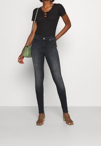 Body negro acanalado con un escote en forma de llave, combinado con jeans skinny de lavado oscuro. Accesorios incluyen un pequeño bolso verde y tacones con tiras.