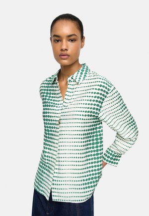 Kobieta w białej koszuli zapinanej na guziki z zielonymi poziomymi wzorami w kropki oraz niebieskich dżinsach, stojąca z jedną ręką za plecami.