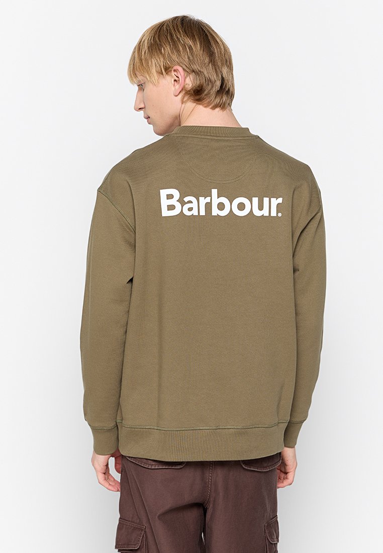 Persona bionda di spalle, che indossa un sudario verde oliva con il logo "Barbour" in bianco sulla schiena e pantaloni cargo marroni.