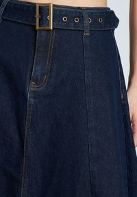 Falda de mezclilla oscura con una cinturilla ancha que presenta una hebilla rectangular dorada y costuras visibles, destacando una textura suave y un diseño acampanado.