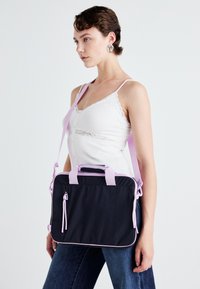 Crna torba od tkanine s ružičastim detaljima, podesivi remen za rame, ručke na vrhu i prednji zip džep. Ima elegantan pravokutni oblik.