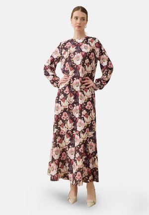 Kvinde stående med hænderne på hofterne iført langærmet, ankel-lang mørk blomsterkjole og beige spidse hæle på hvid baggrund.