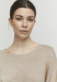 Beige gestricktes Oberteil mit weitem Ausschnitt, subtiler Textur und einem mittigen Nahtdetail. Der Stoff wirkt weich und leicht.