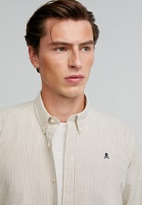 Gestreiftes Button-Down-Hemd in Beige und Weiß mit kleinem marineblauen Logo, klassischem Kragen und strukturiertem Stoff. Weißes Unterhemd sichtbar.