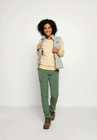 Γκρι fleece γιλέκο πάνω από ένα μπεζ μακρυμάνικο πουκάμισο, σε συνδυασμό με πράσινα παντελόνια και καφέ ορειβατικά μποτάκια, σε στάση μπροστά από έναν ουδέτερο φόντο.