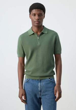 Hombre con una camisa polo verde texturizada de manga corta con cremallera frontal, combinada con vaqueros azules, de pie frente a un fondo liso.