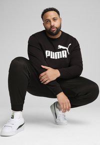 Uomo accovacciato che indossa un felpa nera Puma, pantaloni neri e sneaker bianche Puma su uno sfondo grigio chiaro uniforme.