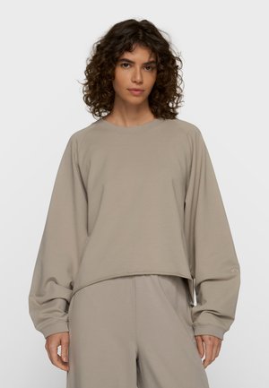 Kvinde med krøllet hår iført en løs, beige sweatshirt og matchende bukser, stående mod en ensfarvet lys baggrund.