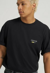 Sort bomulds T-shirt med oprevne ærmer, der har et lille broderet logo i hvidt og gult på venstre brystområde. Ensartet struktur.