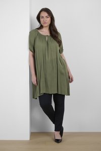 Tunique verte olive avec décolleté en scoop et détail en forme de trou de serrure, associée à un jeans skinny noir et des chaussures noires à talons pointus, sur un fond neutre.