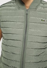 Olive green puffer vest with horizontal quilted pattern, zip closure, ribbed collar, and embroidered crocodile logo on the left chest.