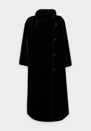 Žena nosiaca čierny oversized kabát, sivý rolák, široké čierne nohavice a malé okrúhle slnečné okuliare stojaca na jednoduchom pozadí.