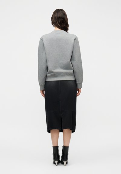 Γυναίκα με σκούρα μαλλιά που φοράει γκρι φούτερ, μαύρη μίντι φούστα με σκίσιμο στο πίσω μέρος και μαύρα μποτάκια μέχρι τον αστράγαλο, στέκεται με την πλάτη σε απλό φόντο.