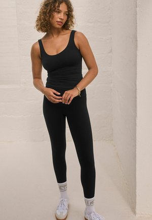 Schwarzes, figurbetontes Tanktop und Leggings aus dehnbarem Material. Das Model trägt weiße Sneakers mit gemusterten Socken vor einem hellen Hintergrund.