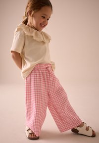 Cămașă de culoare bej deschis cu mâneci scurte și guler dantelat cu volane, combinată cu pantaloni largi în carouri roz și albi și sandale albe.