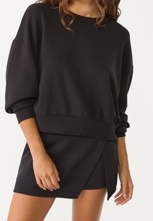 Kvinde iført en sort langærmet sweatshirt og matchende sort mininederdel med slå-om-design, stående med den ene hånd tæt ved hoften.